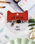 A colourful cat collar with a breakaway safety clasp, displayed on a card shaped like a cat's head, with the text 'CAT COLLAR' and 'ANDY PAWHOL'. The collar is laid out on a desk with various desk accessories.