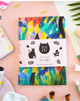 A colourful tea towel with a pattern that includes a silhouette of a black cat and the text 'NIASKI cats in art tea towel'. The tea towel is displayed with various art supplies around it, such as paint tubes and brushes.