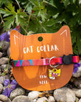 Cat collar with colorful straps and charms on an orange card labeled 'Cat Collar' by Paw Klee, set against a natural background with rocks and plants.