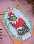 Decorative card with a cat wearing a Santa outfit and Christmas decorations on a pink background.