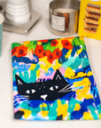 Colourful cat tea towel gift with a black cat design on a table with various items.