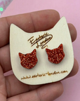 Cat-shaped earrings with glittery details on a wooden card held by a hand against a pink background.
