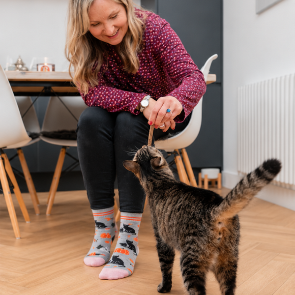 A woman in a pink polka-dot sweater and patterned socks pets a tabby cat on a light wood floor in a modern living room setting.