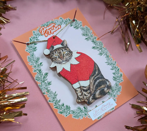 Decorative card with a cat wearing a Santa outfit and Christmas decorations on a pink background.