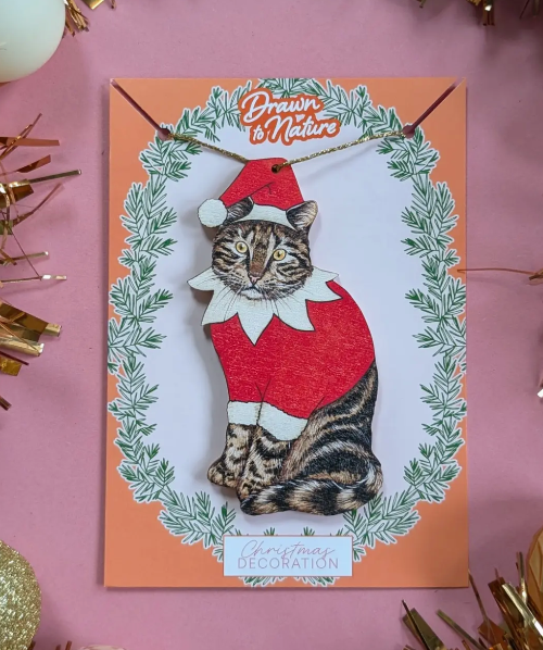Decorative card with a cat in an elf costume surrounded by greenery on a pink background.