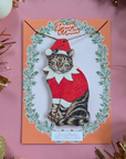 Decorative card with a cat in an elf costume surrounded by greenery on a pink background.