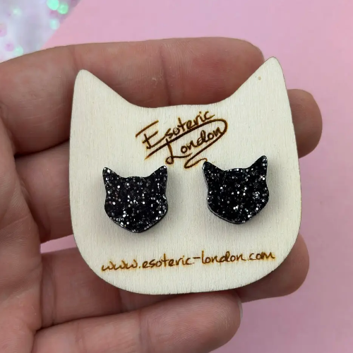 Black glitter cat ear earrings on a white card with &#39;Esoteric London&#39; branding, held by a hand against a pink background.