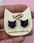 Black glitter cat ear earrings on a white card with 'Esoteric London' branding, held by a hand against a pink background.