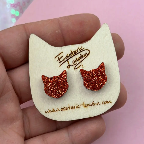Cat-shaped earrings with glittery details on a wooden card held by a hand against a pink background.
